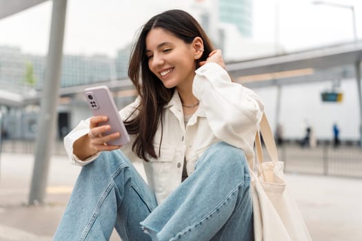 Bright portrait of cheerful young woman 25s with shopper on shoulder waiting public transport at station smiling beautifully and texting.