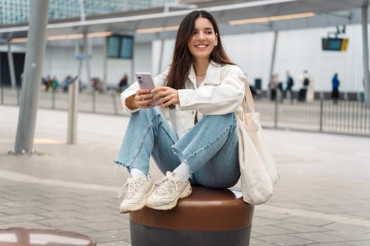 Bright portrait of cheerful young woman 25s with shopper on shoulder waiting public transport at station smiling beautifully and texting.