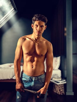 Sexy handsome young man standing shirtless in his bedroom under a window, looking at camera