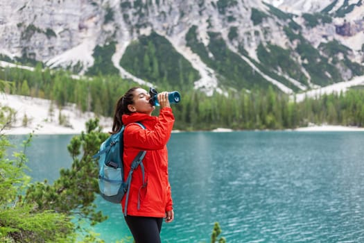 Hydration while on track. Attractive woman hiker in red raincoat drinking water near a lake with mountains in the background.