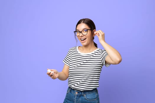 attractive brunette in striped t-shirt using small white wireless headphones against purple very peri color background.