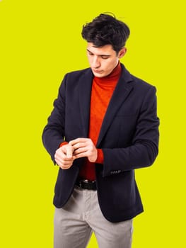 Handsome young man in studio, adjusting his jacket, looking at sleeve with serious expression, wearing turtleneck sweater on yellow background