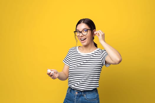Funny attractive multi ethnicity young woman with glasses using in-ears wireless headphones against yellow background.