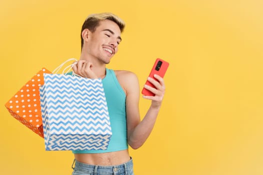 Happy gay man using the mobile while holing shopping bags in studio with yellow background