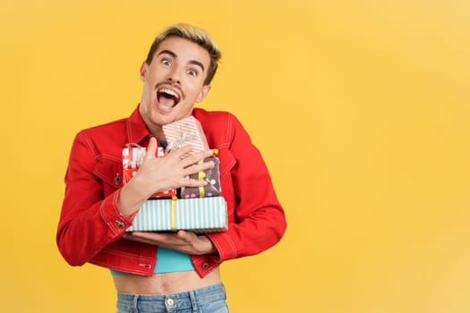 Happy gay man holing many colorful gifts in studio with yellow background