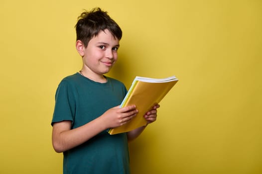Handsome Caucasian teenage boy 10-12 years old in green casual t-shirt, smiling looking at camera, holding textbooks, ready to start a new semester of academic school year, isolated yellow background