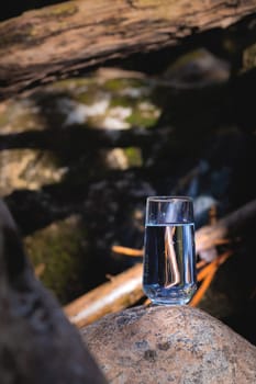 Pure water in a transparent glass cup stands on a stone next to a stormy mountain stream in a coniferous forest. Clean water concept.