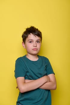 Emotional portrait of a sorrowful, overwhelmed school boy in green t-shirt, expressing sad emotions, looking at camera, posing with arms folded, isolated on yellow color background with copy ad space.
