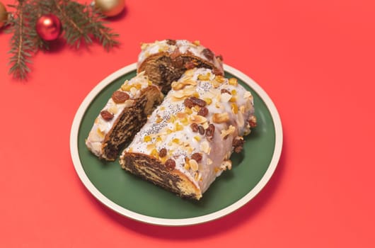 Christmas strudel with poppy seeds, traditional polish festive pie Makowiec on green plate,covered with sugar icing,walnuts,dried fruits on red background with Christmas tree, toy balls. Horizontal.