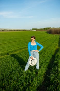 A cute woman in jeans and a hat stands in a green field. A smiling woman in a blue top and jeans walks in the green grass.