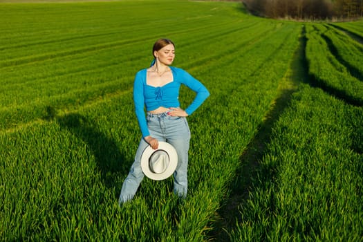 A cute woman in jeans and a hat stands in a green field. A smiling woman in a blue top and jeans walks in the green grass.