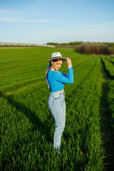 A cute woman in jeans and a hat stands in a green field. A smiling woman in a blue top and jeans walks in the green grass.