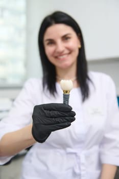 Blurred smiling female dentist orthodontist surgeon holds a realistic model of enlarged human tooth that shows the mechanism of dental implant installation. Denture. Prosthesis. Maxillofacial surgery