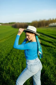 A cute woman in jeans and a hat stands in a green field. A smiling woman in a blue top and jeans walks in the green grass.