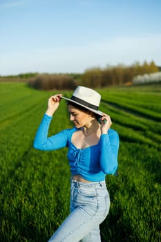 A cute woman in jeans and a hat stands in a green field. A smiling woman in a blue top and jeans walks in the green grass.