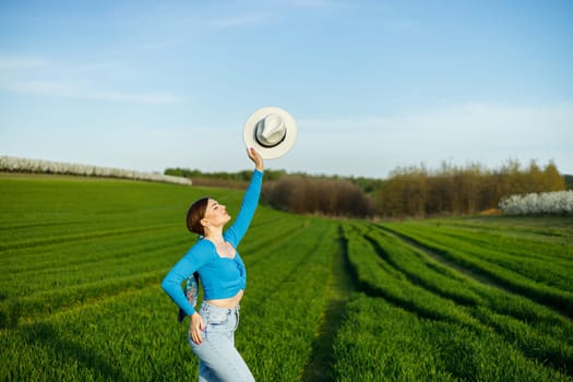 Young attractive woman in white hat, blue shirt, blue jeans, posing in summer green field. Copy, empty space for text