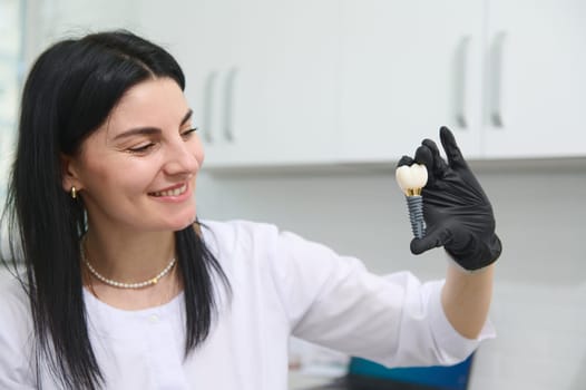 Beautiful woman dentist, orthodontist, maxillofacial surgeon smiles, showing an enlarged model of a human tooth to the camera, explaining the mechanism of installing a dental implant. Dental practice