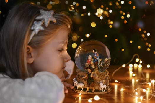 Girl looking at a glass ball with a scene of the birth of Jesus Christ near christmas tree