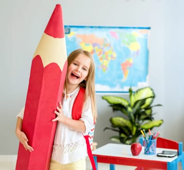 Little smiling blond girl with red bag holding huge red decorative pencil on her shoulder in the school classroom