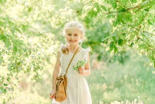 Cute little girl is walking between the trees, holding bag and flower, in green park