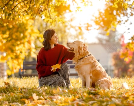 Beautiful girl with golden retriever dog sitting in autumn park with yellow leaves. Pretty young woman petting purebred doggy labrador at fall season at nature
