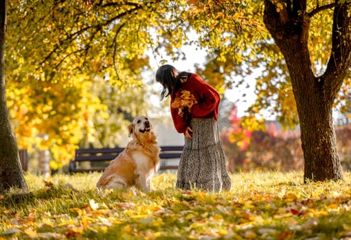 Beautiful girl with golden retriever dog holding yellow leaf in hand in autumn park. Pretty young woman playing with purebred doggy labrador at fall season at nature