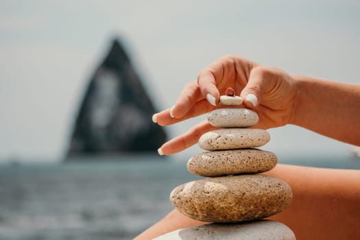 Woman bilds stones pyramid on the seashore on a sunny day on the blue sea background. Happy holidays. Pebble beach, calm sea, travel destination. Concept of happy vacation on the sea, meditation, spa