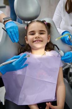 Top view. Hands of a dentist and assistant hold sterile dental instruments - dental mirror, probe, drill and saliva ejector above a smiling little patient at appointment in pediatric dentistry clinic