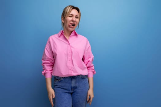 young energetic European blond leader woman with ponytail dressed in a pink blouse makes a grimace against the background with copy space.