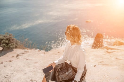 Business woman on nature in white shirt and black skirt. She works with an iPad in the open air with a beautiful view of the sea. The concept of remote work