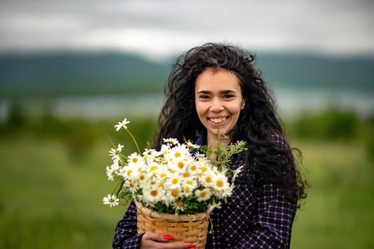 A woman stands on a green field and holds a basket with a large bouquet of daisies in her hands. In the background are mountains and a lake