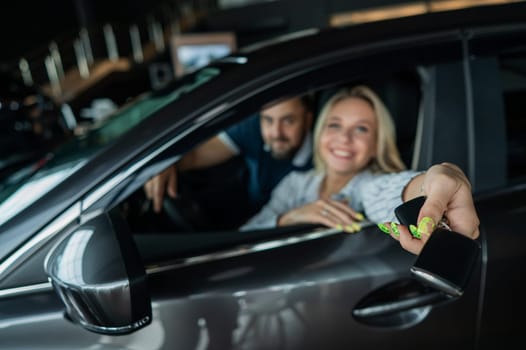Happy caucasian couple is sitting in a car in a car dealership. A woman holds the keys to a new car
