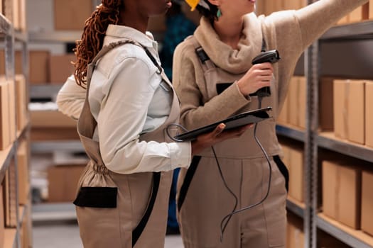 Storehouse colleagues reaching for cardboard box on shelf to scan barcode. Warehouse operators managing received parcels registration with bar code scanner and digital tablet software