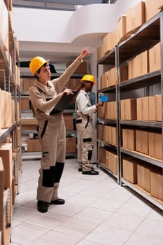 Storage employees coordinating shipping process and preparing boxes before transportation. Warehouse manager wearing jumpsuit and protective helmet, holding clipboard and pointing at cartons