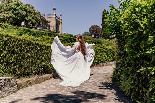 Brunette runs white dress park. A beautiful woman with long brown hair and a long white dress runs along the path along the beautiful bushes in the park, rear view.