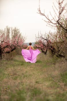 Woman peach blossom. Happy curly woman in pink dress walking in the garden of blossoming peach trees in spring.