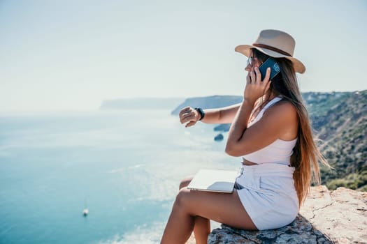 Successful business woman in yellow hat working on laptop by the sea. Pretty lady typing on computer at summer day outdoors. Freelance, travel and holidays concept.