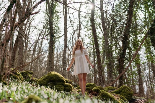 Snowdrops galanthus blonde. A girl in a white dress sits on a meadow with snowdrops in a spring forest.