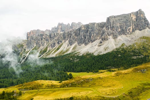 Rocky Dolomite Alps peak covered by clouds and green forest at the foot of the mountain.