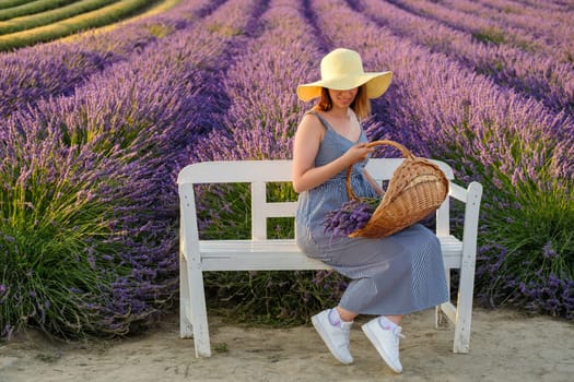 Young woman donning a straw hat and sundress sits on the bench nestled in a vibrant lavender field and holding a basket with flowers
