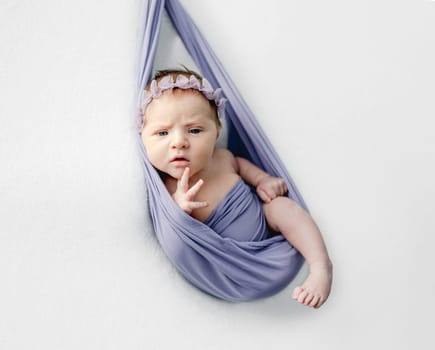 Newborn baby girl with beautiful blue eyes in hammock studio portrait. Cute infant child kid wearing wreath looking at camera