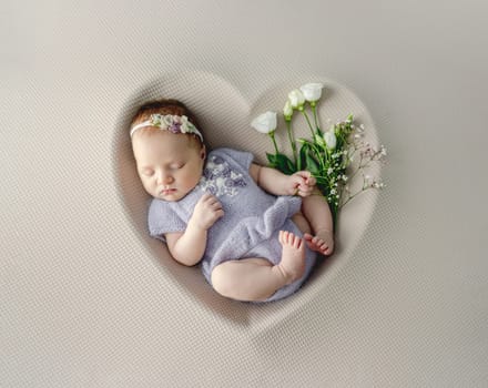 Newborn baby girl wearing knitted dress and wreath sleeping in heart shape with white roses studio portrait.