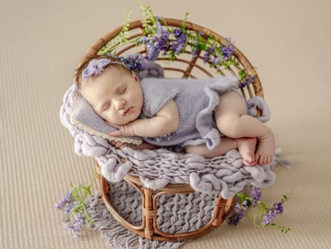 Newborn baby girl wearing knitted dress and wreath sleeping on chair decorated with purple flowers. Cute infant child kid napping