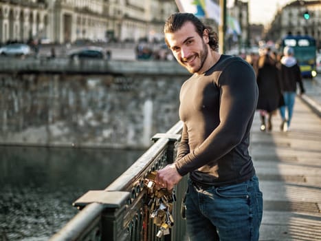 Side view of bearded man in casual clothes standing on footbridge and attaching love lock to metal railing and smiling to camera