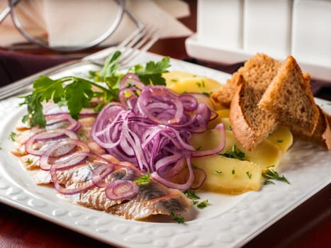 Appetizer of salted herring, red onion, potatoes and rye bread in white plate on dark brown wooden table in restaurant