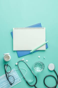 Drugs and glass of water on green medical background top view. Stethoscope and notebook with a pen at the workplace of a doctor. Nurse desktop concept. Vertical photo.