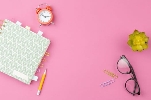 Spiral notebook with foliage ornament, bookmarks from paper clips and sheets for notes, glasses, pencil, alarm clock and a flower in a pot on a pink background. Top view.