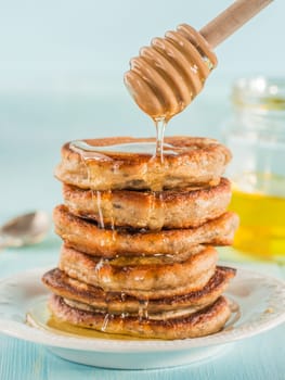 Stack of small pancakes with banana and chia seeds in honey on light blue background. Honey pouring over stack of mini pancakes with chia or poppy seeds.