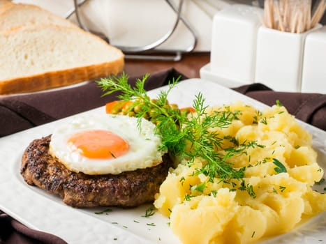 Beefsteak with fried egg and mashed potatoes on white plate close-up on dark brown wooden table in restaurant