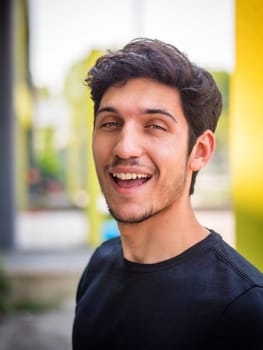 Head and shoulders shot of one handsome young man with green eyes laughing, in urban setting, looking at camera, wearing t-shirt